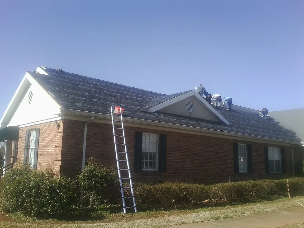 Crew tearing shingles off a brick ranch roof with a ladder set — Metal Roof Repair work by our crew in Acme