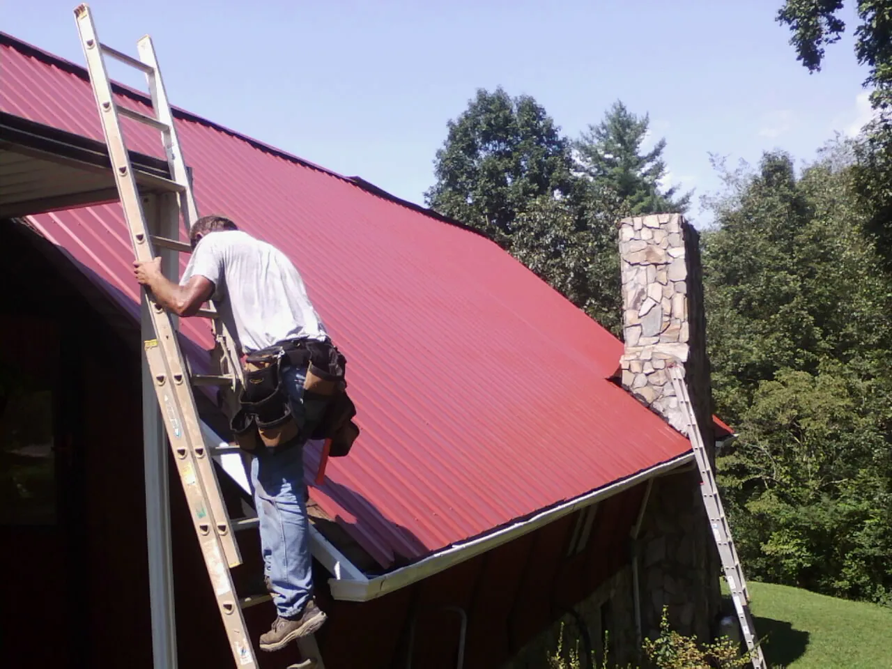 Roofer on a ladder beside a red metal roof and stone chimney — Soffit & Fascia Repair by Roofing Brothers in Acme